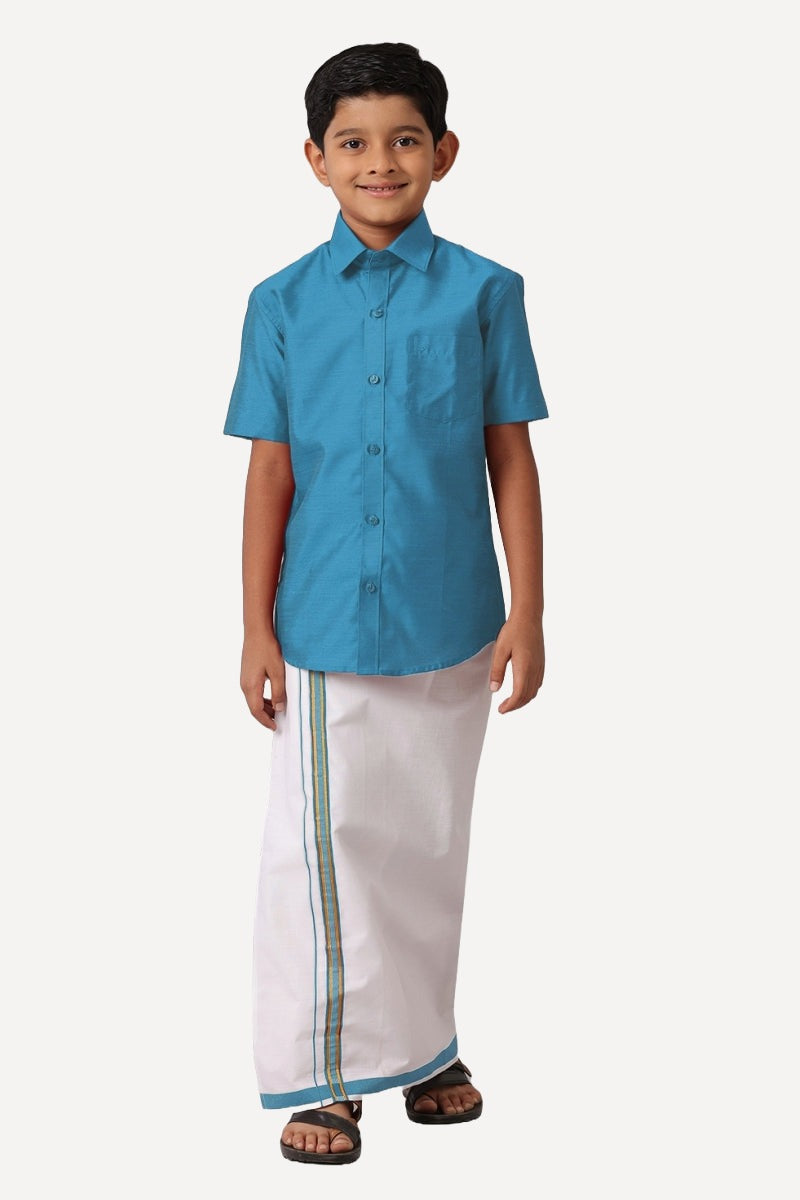A boy wearing a blue shirt and white dhoti with blue trimmings standing against a white background.