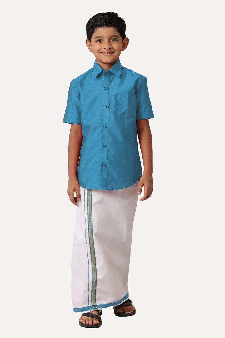 A boy wearing a blue shirt and white dhoti with blue trimmings standing against a white background.