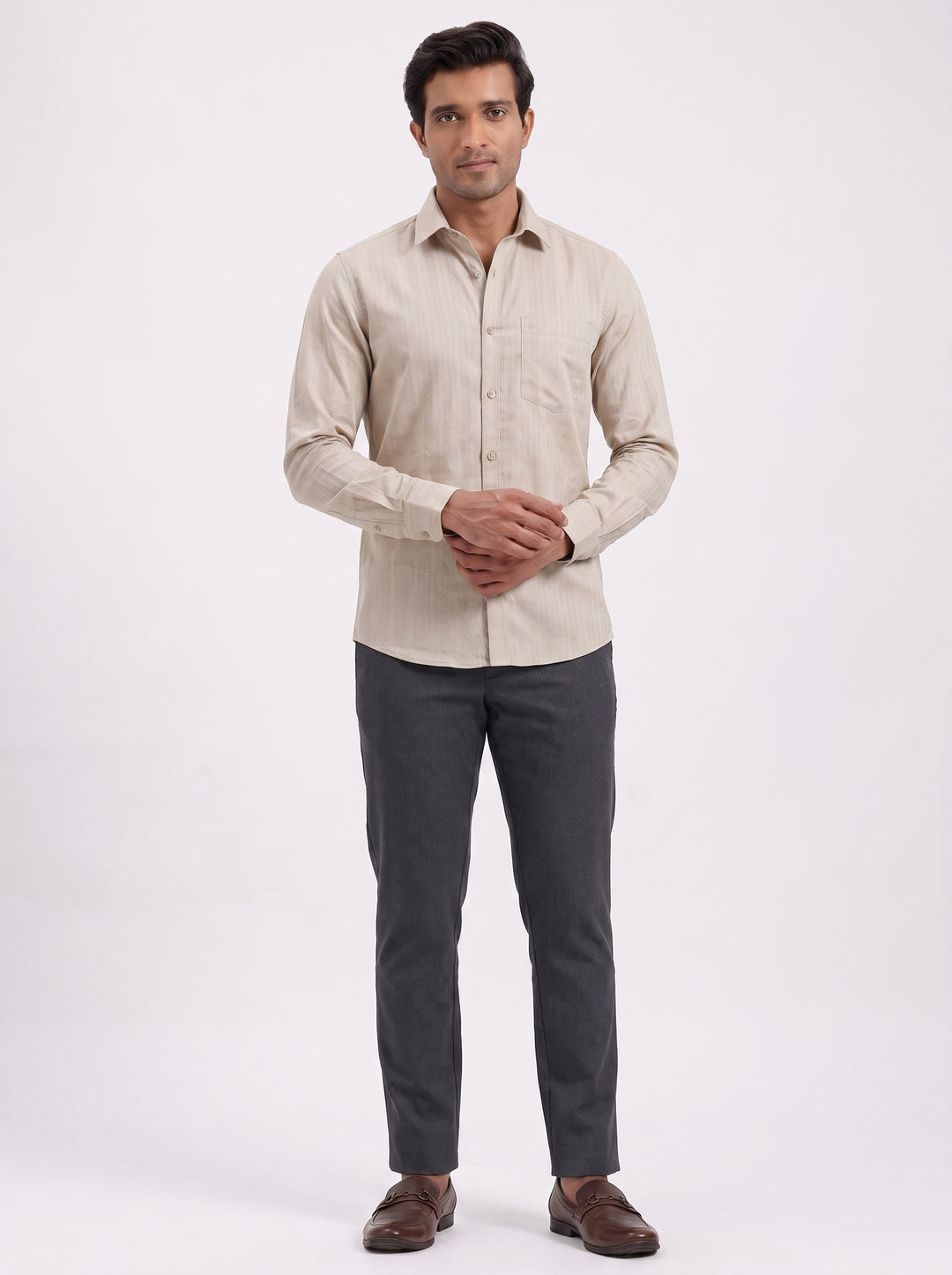 A man modeling a light beige long-sleeve linen shirt paired with dark gray trousers and brown leather shoes, standing against a plain white studio background.