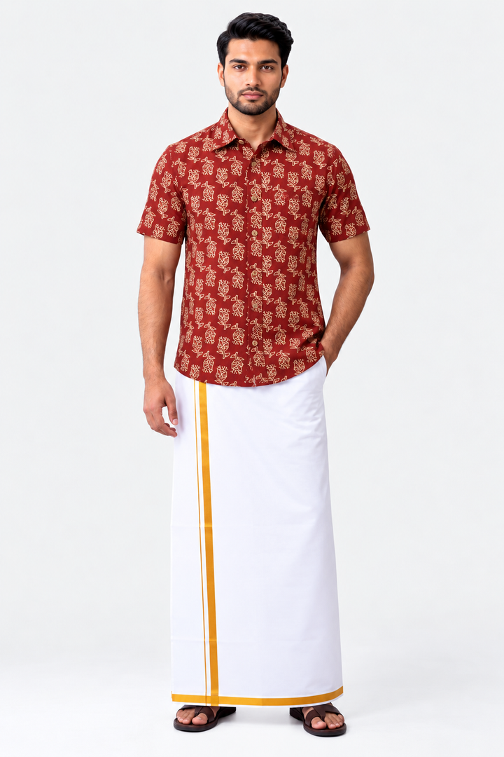 A man modeling a traditional Indian outfit consisting of a rust-red short-sleeve button-up shirt with a beige floral pattern and a white drape (kani) with a yellow border, paired with black sandals, against a plain white studio background.