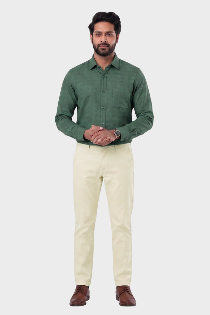 A man modeling a dark green long-sleeve button-up shirt paired with cream-colored trousers and brown leather shoes, standing against a solid light gray background.