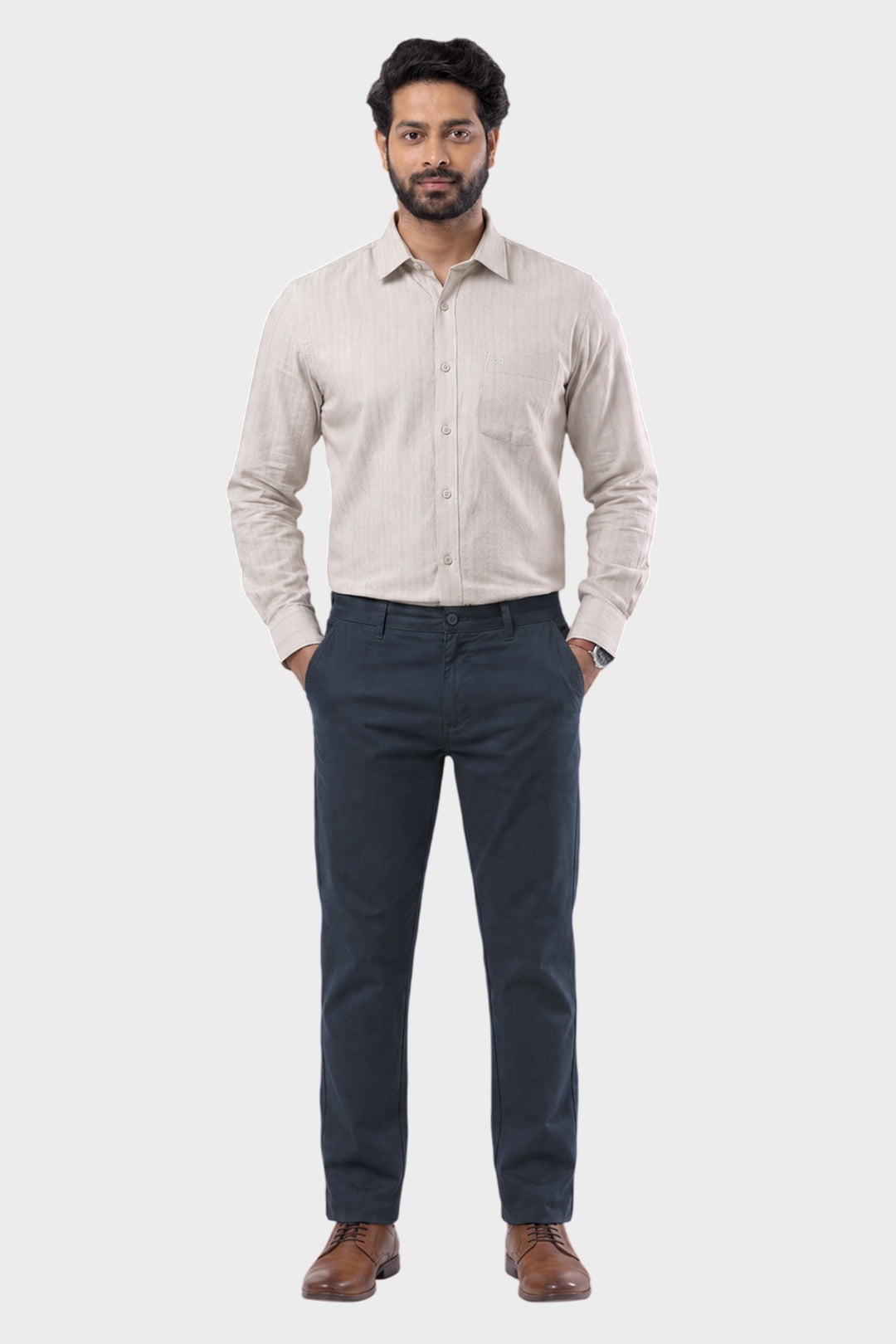 A man wearing a light beige long-sleeve button-down shirt and navy blue trousers, standing against a solid light gray background.