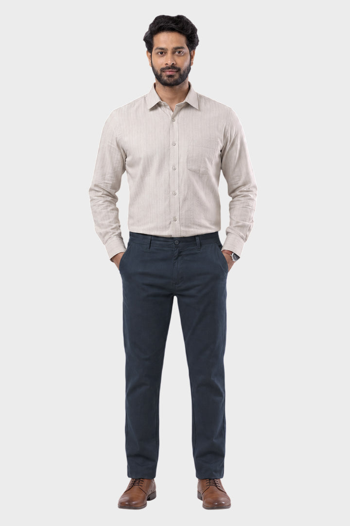 A man wearing a light beige long-sleeve button-down shirt and navy blue trousers, standing against a solid light gray background.