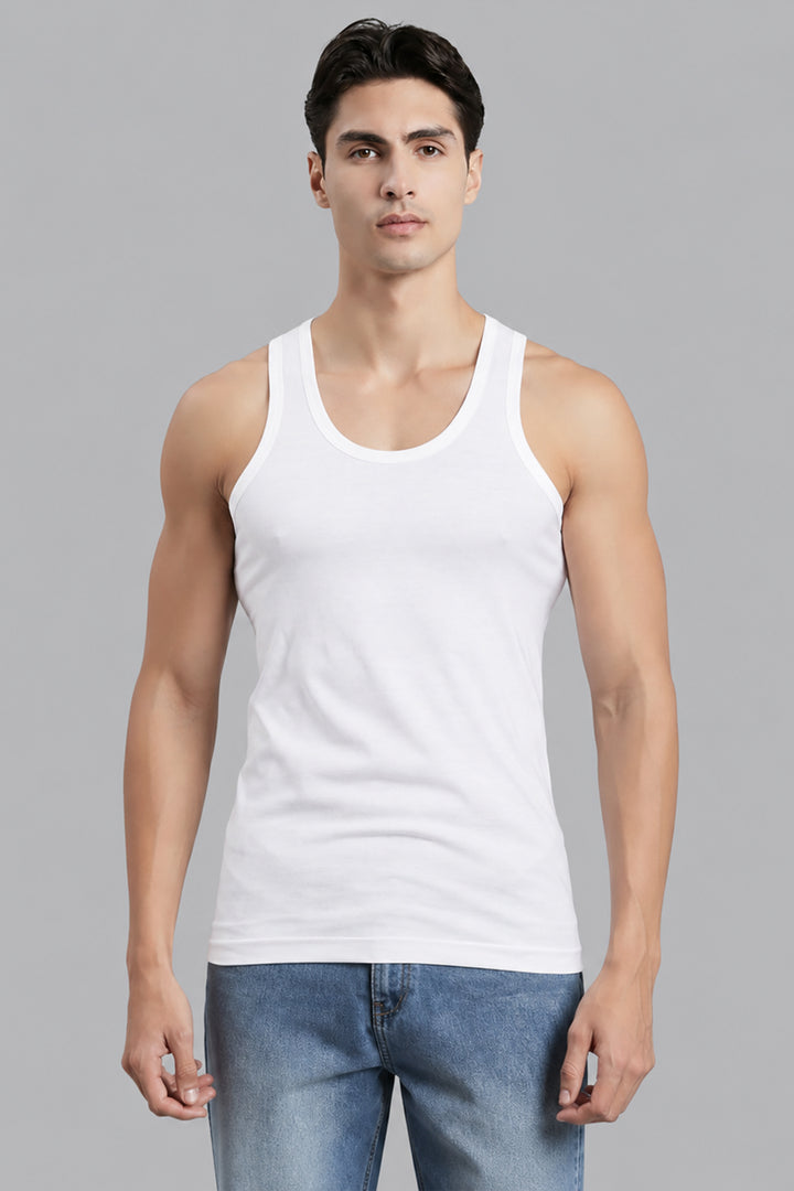 A man wearing a white ribbed tank top and blue jeans, standing against a solid gray studio background.
