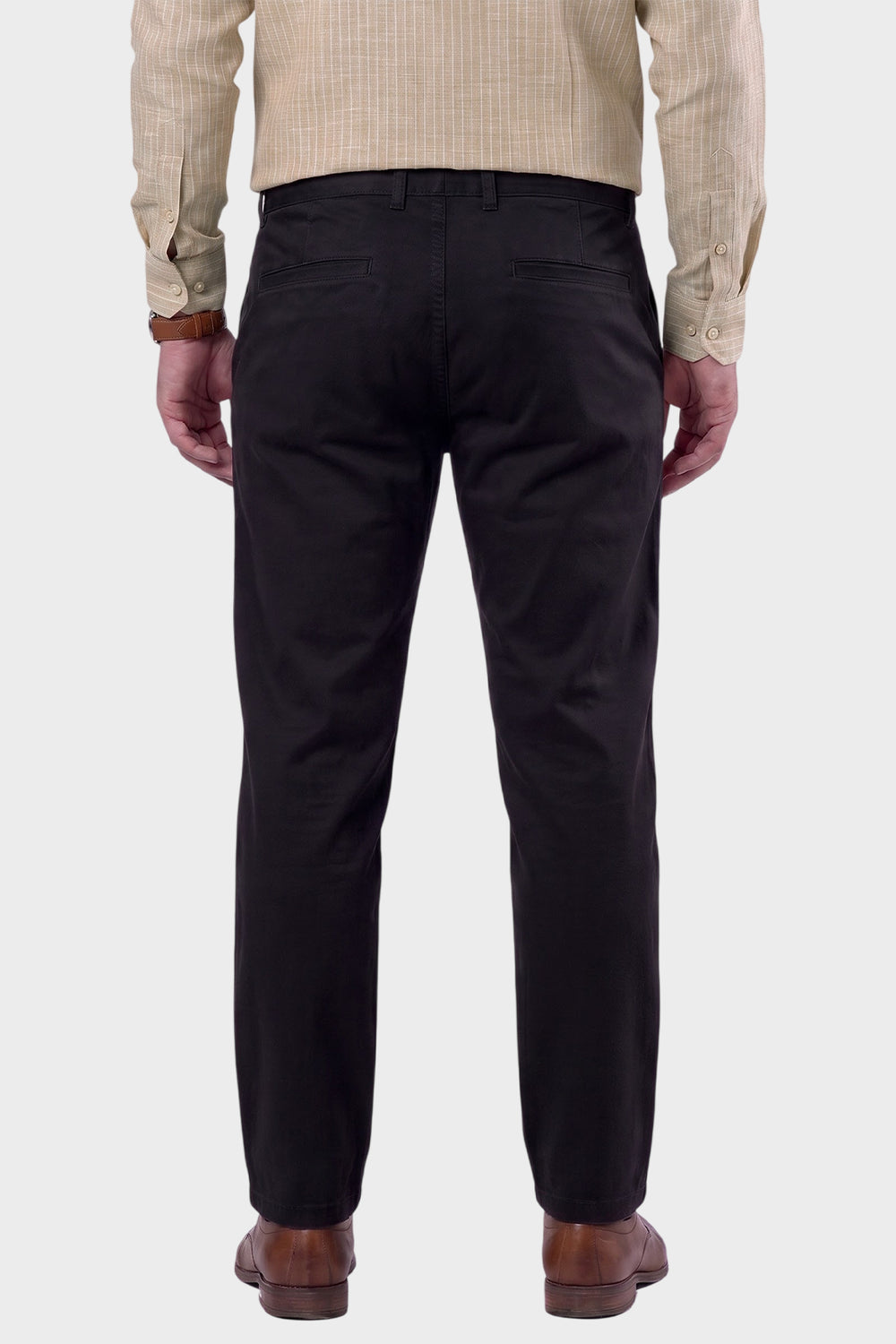 Rear view of a man wearing black trousers, a beige checkered shirt, and brown leather shoes, standing against a light gray background.