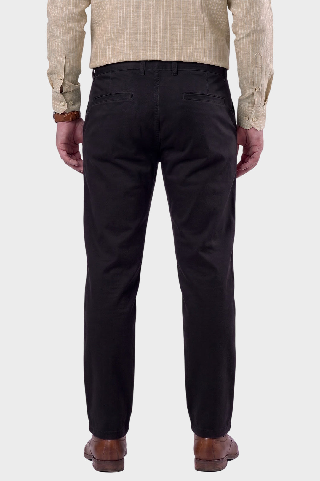 Rear view of a man wearing black trousers, a beige checkered shirt, and brown leather shoes, standing against a light gray background.