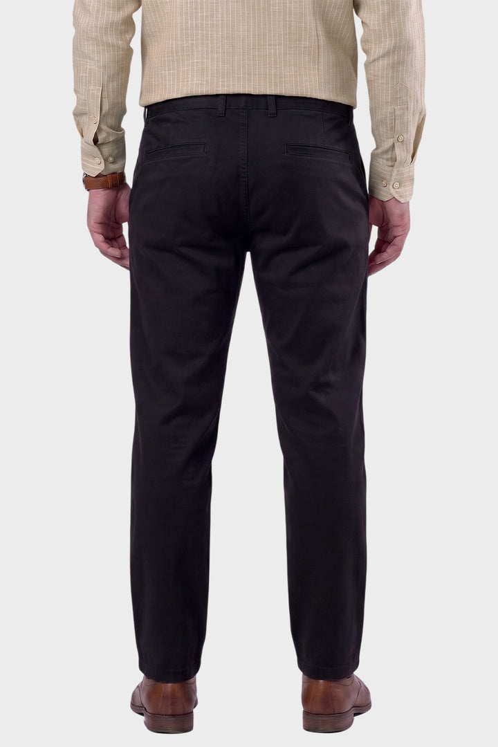 Rear view of a man wearing black trousers, a beige checkered shirt, and brown leather shoes, standing against a light gray background.