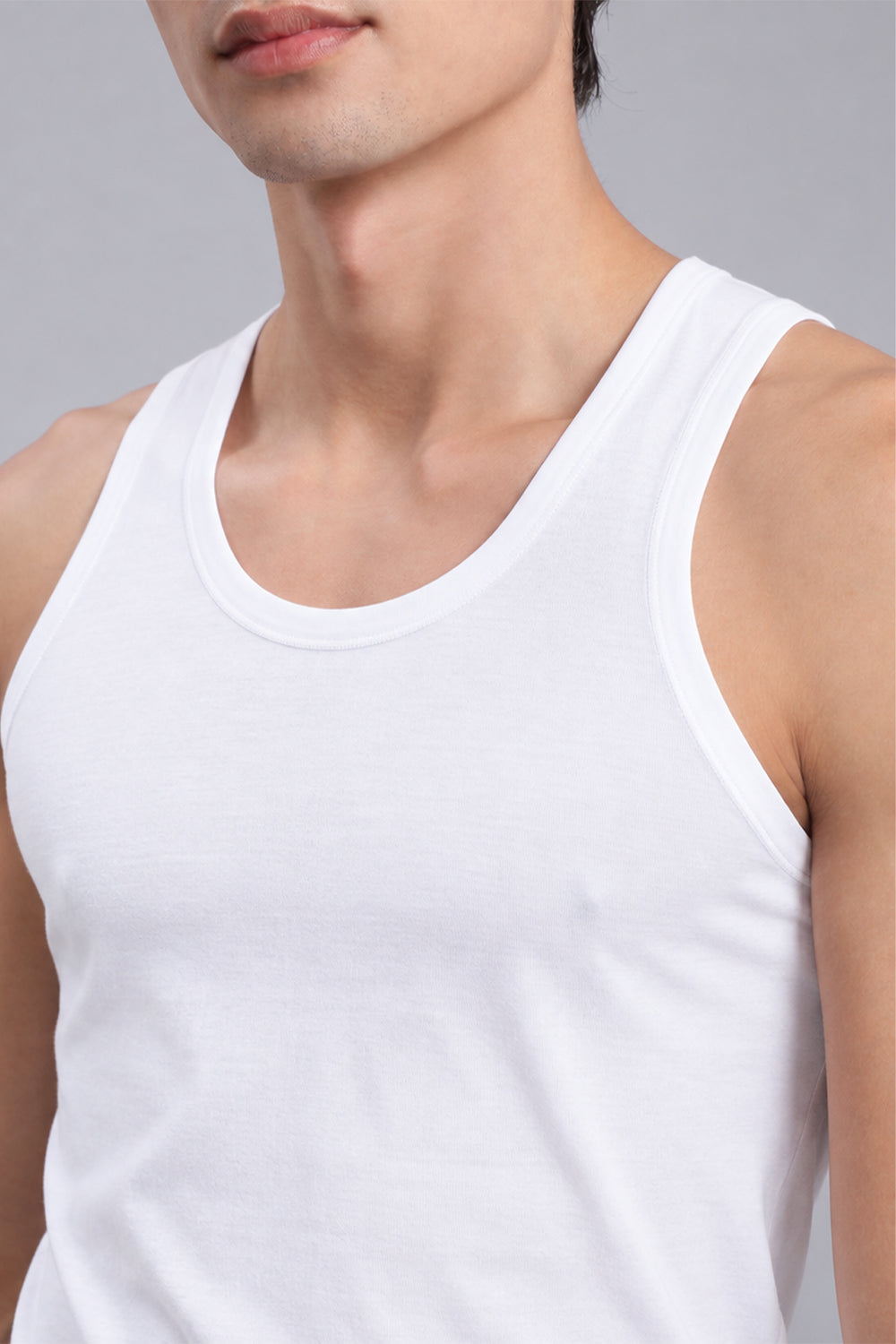 A close-up of a man wearing a plain white ribbed Vest top against a light gray studio background.