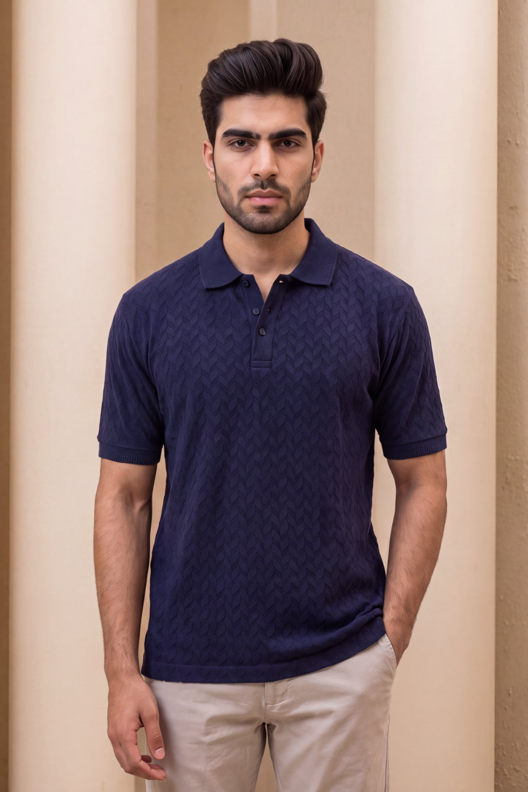 A man wearing a dark navy blue short-sleeve polo shirt with a subtle textured chevron pattern, paired with beige trousers, standing in front of a neutral beige background.