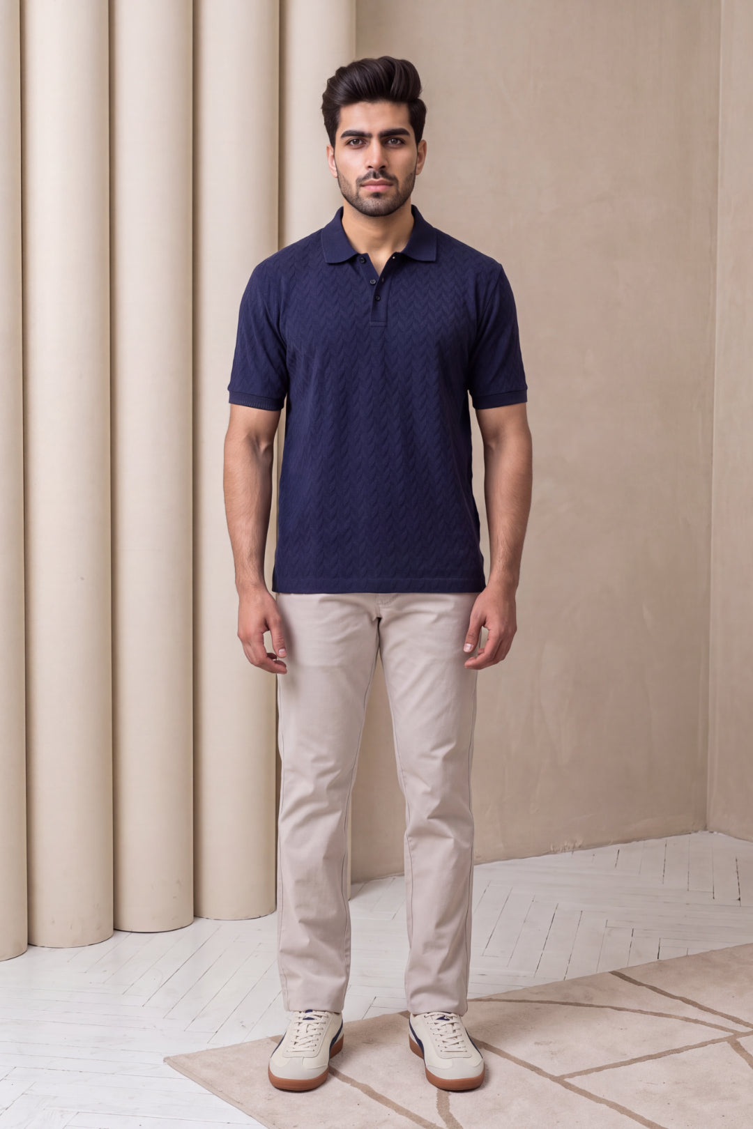 A man wearing a navy blue short-sleeve polo shirt with a subtle textured pattern, paired with beige trousers and white sneakers, standing in front of a beige wall with decorative columns.