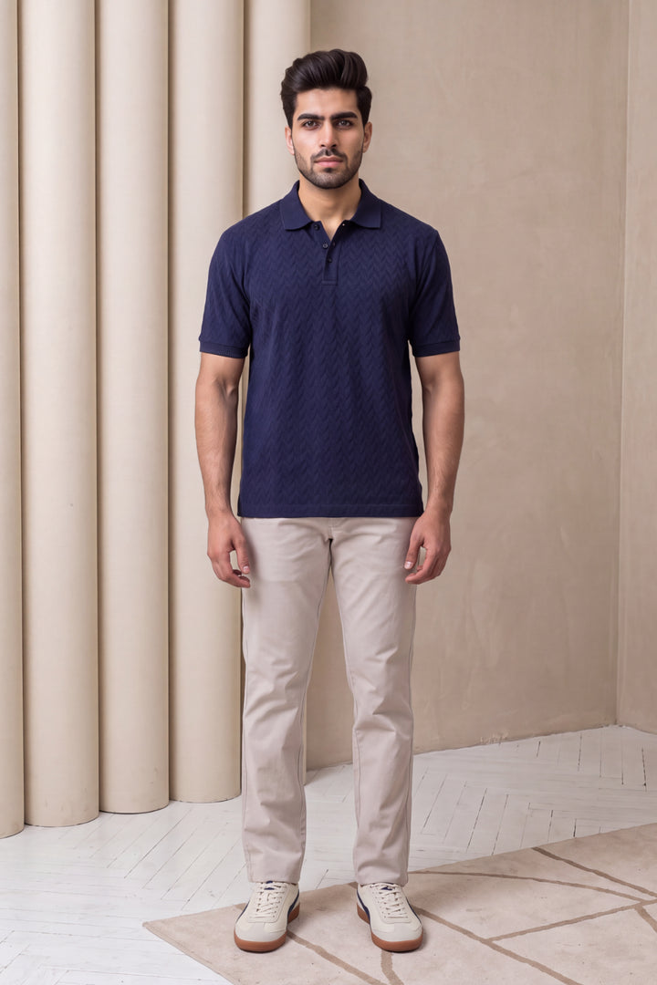 A man wearing a navy blue short-sleeve polo shirt with a subtle textured pattern, paired with beige trousers and white sneakers, standing in front of a beige wall with decorative columns.