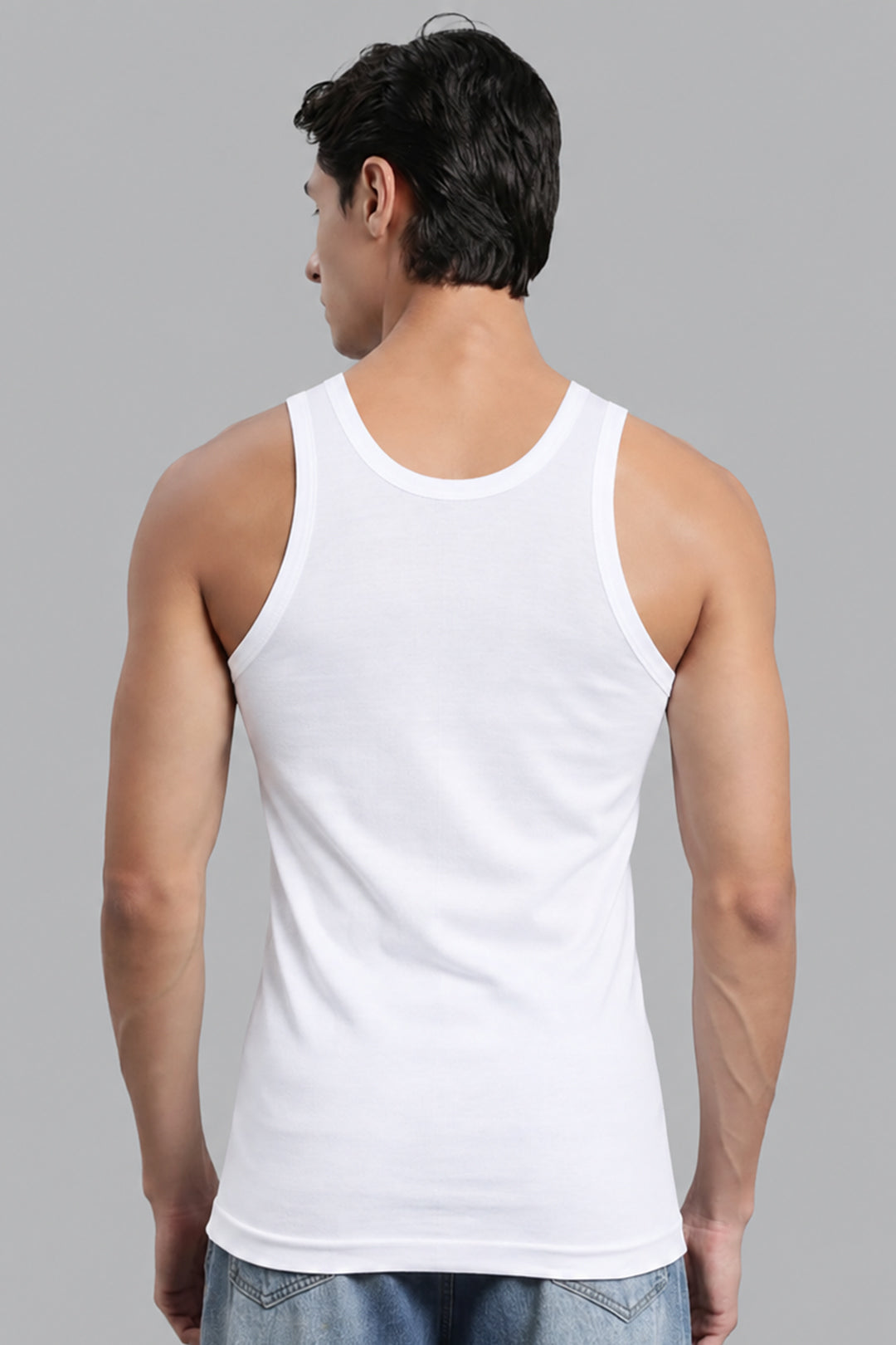 A man viewed from the back wearing a plain white ribbed Vest top and light blue jeans against a solid gray studio background.