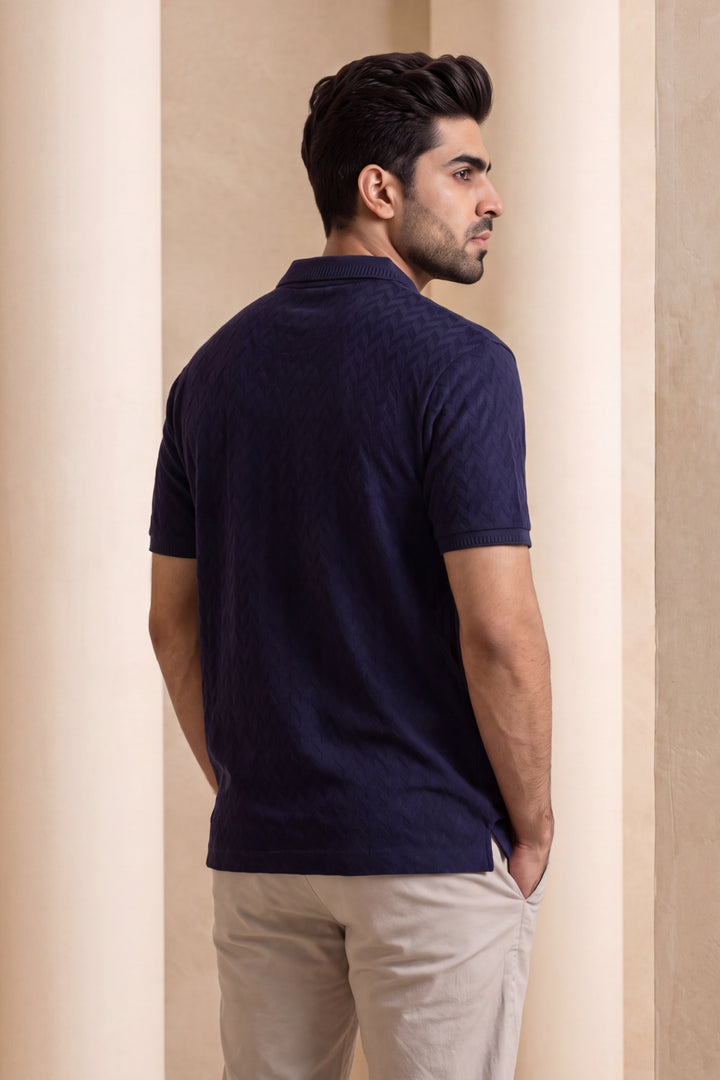 A man wearing a dark navy blue textured short-sleeve polo shirt and beige trousers, standing in profile against a neutral beige architectural background.