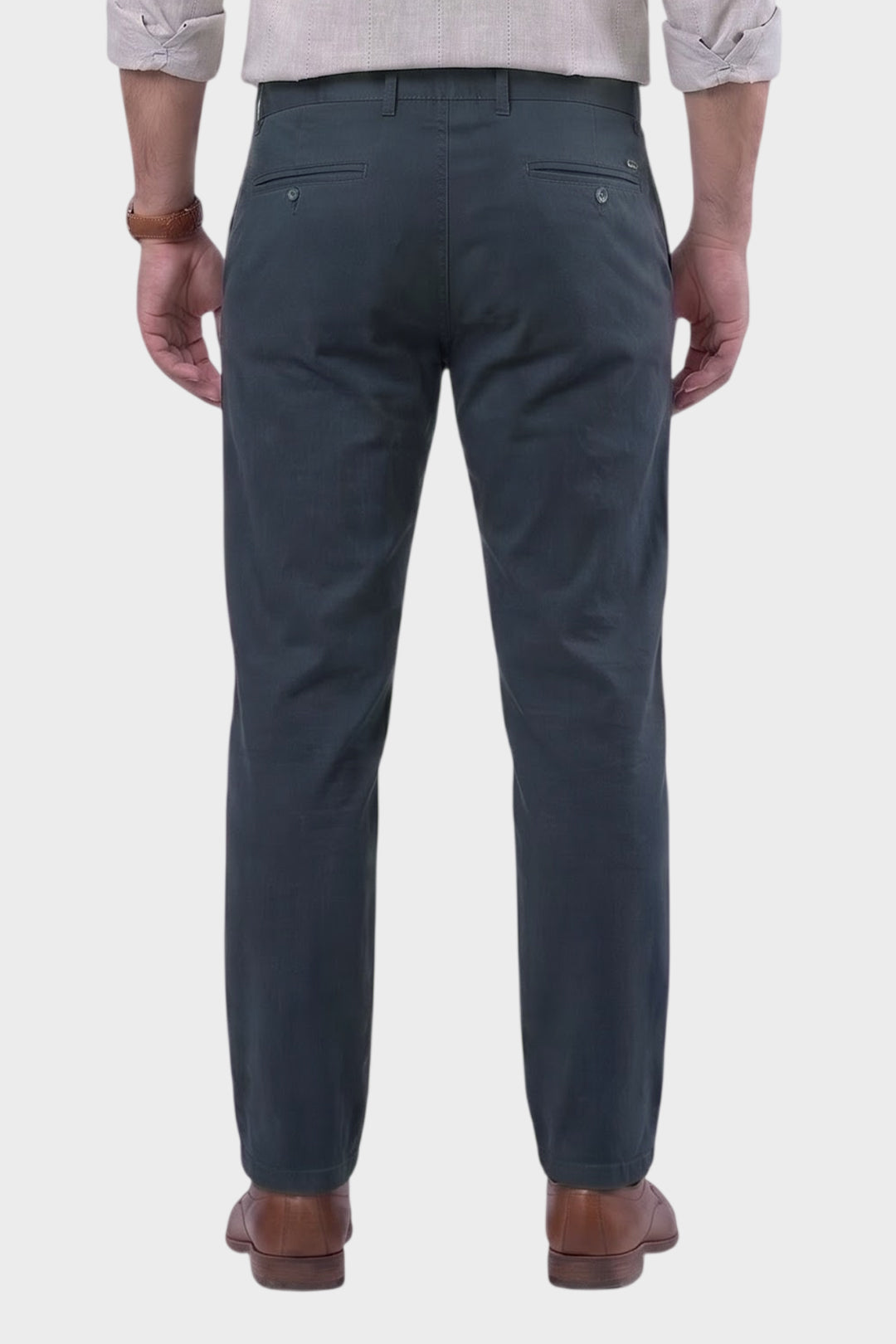 Rear view of a man wearing dark slate blue chinos with brown leather shoes, shown against a light gray background.