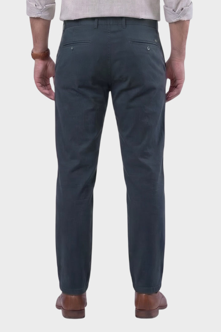 Rear view of a man wearing dark slate blue chinos with brown leather shoes, shown against a light gray background.