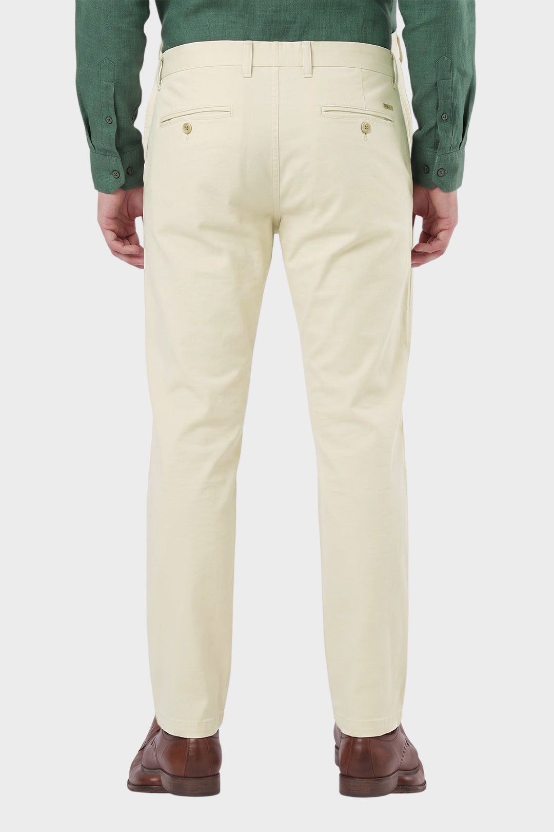 A man viewed from the back wearing light cream-colored chino pants, a green long-sleeve shirt, and brown leather shoes, set against a plain light gray studio background.