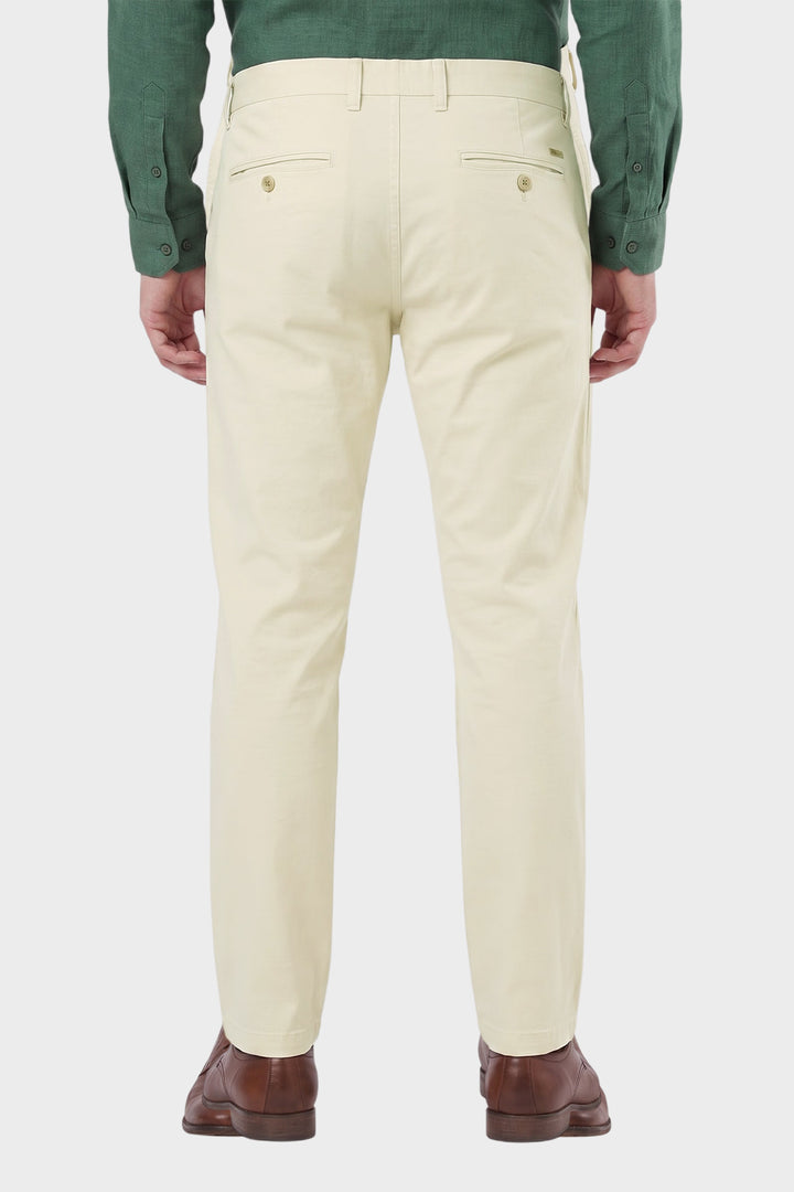 A man viewed from the back wearing light cream-colored chino pants, a green long-sleeve shirt, and brown leather shoes, set against a plain light gray studio background.