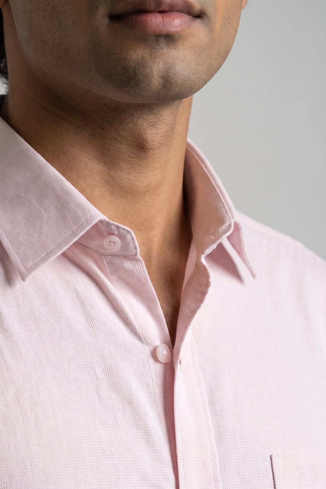 Close-up of a man wearing a light pink textured button-down shirt, showing the collar, button, and chest area against a neutral gray background.