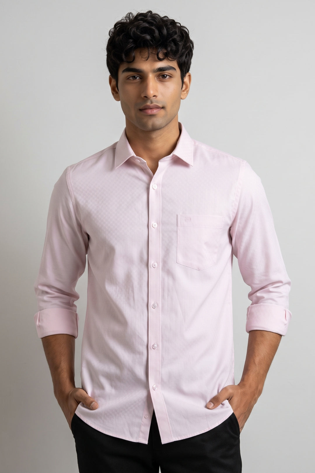A man wearing a pink long-sleeve button-down shirt with a subtle textured pattern, paired with black trousers, standing against a neutral light gray background.