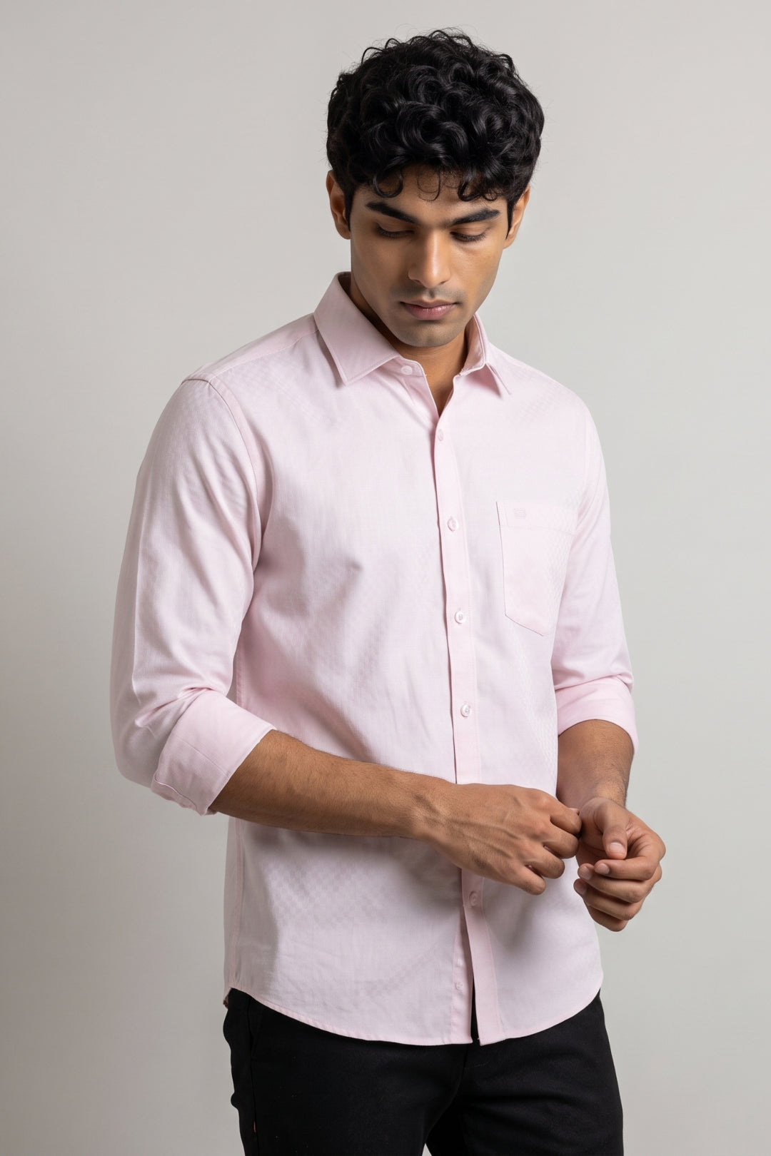 A man wearing a light pink long-sleeve button-down shirt with black pants, standing against a plain light gray studio background.
