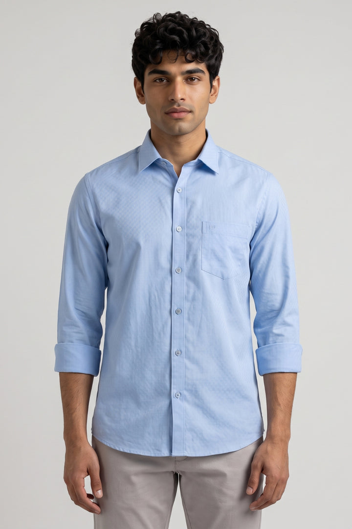 A man wearing a light blue long-sleeve button-down shirt with a subtle textured pattern, paired with light gray trousers, standing against a neutral light gray background.