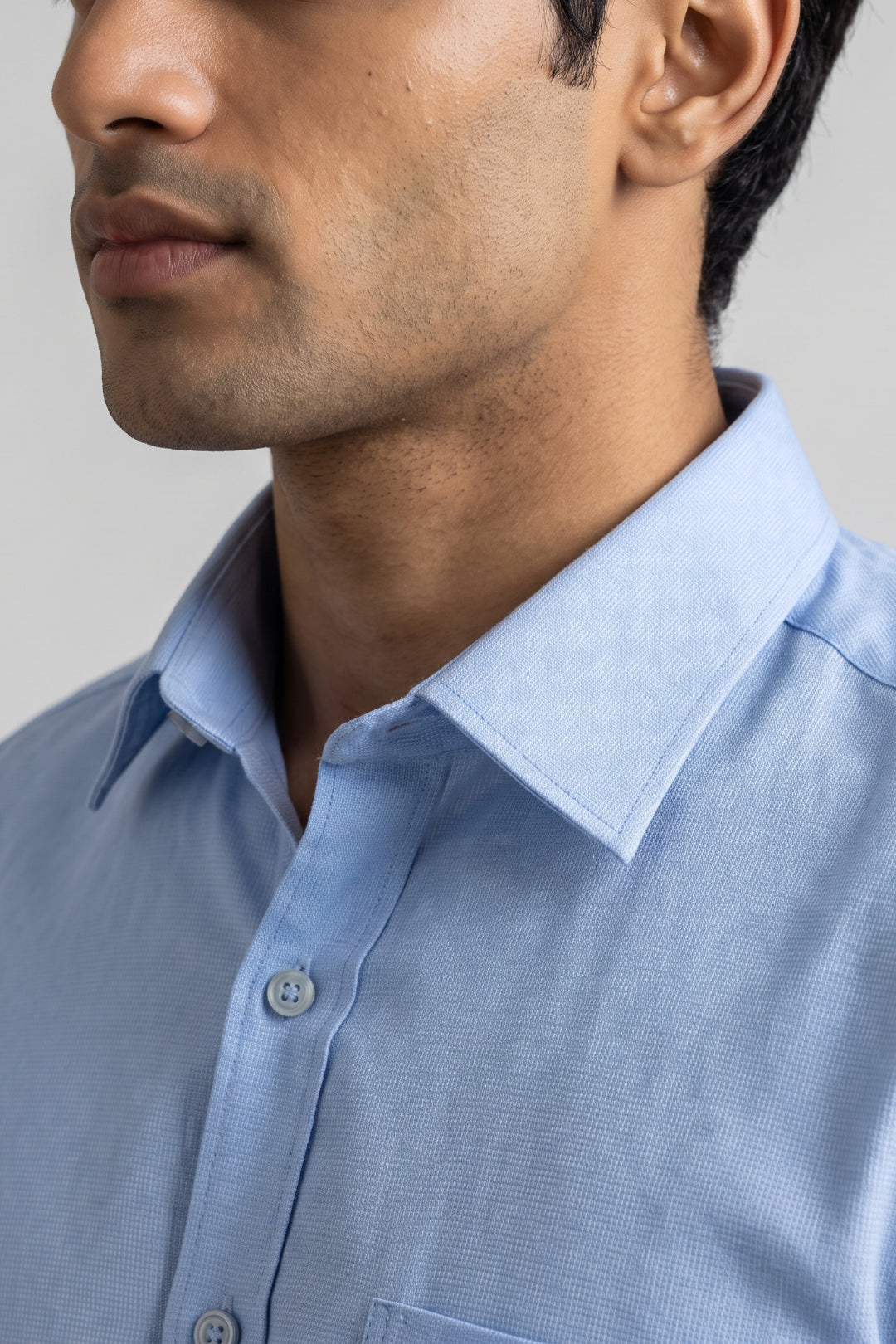 Close-up of a man wearing a light blue textured dress shirt, showing the collar, button placket, and chest pocket area against a neutral gray background.