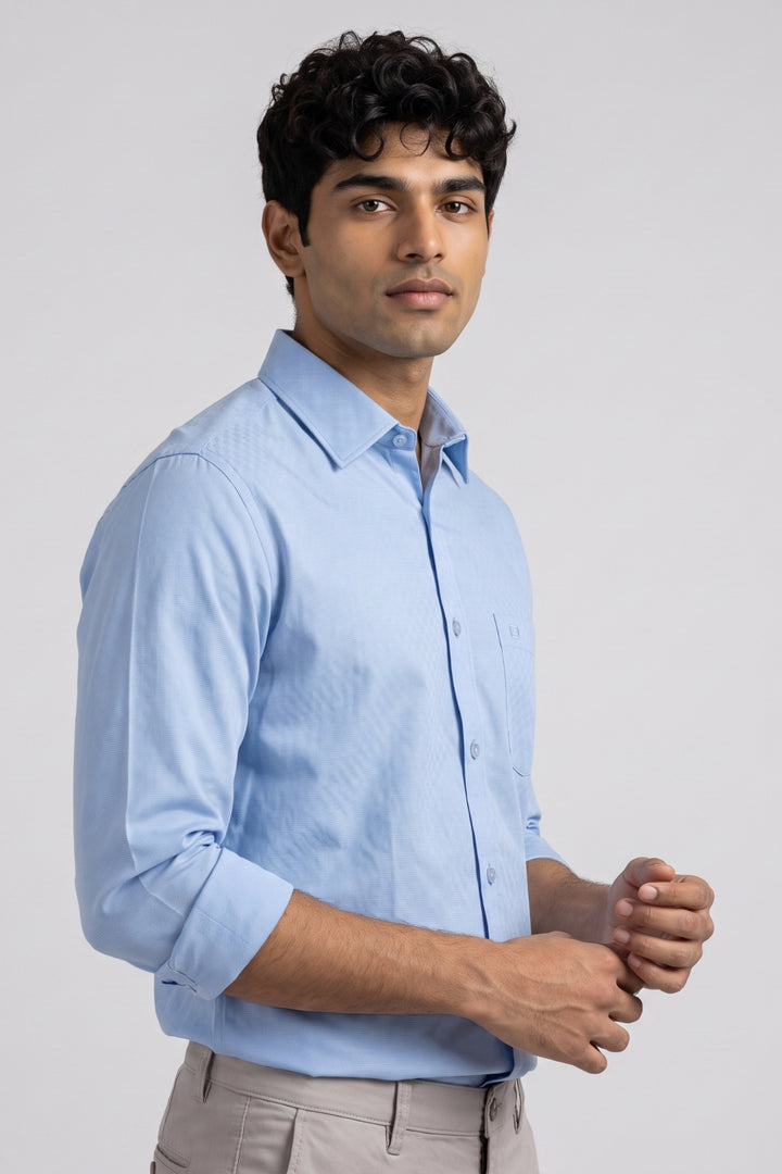 A man wearing a light blue long-sleeve button-down shirt and beige trousers, standing against a light gray studio background.