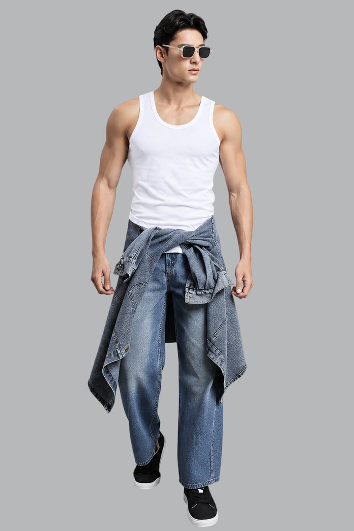 A muscular man wearing aviator sunglasses, a white Vest top, blue jeans, and a tied blue denim jacket, walking against a solid gray background.