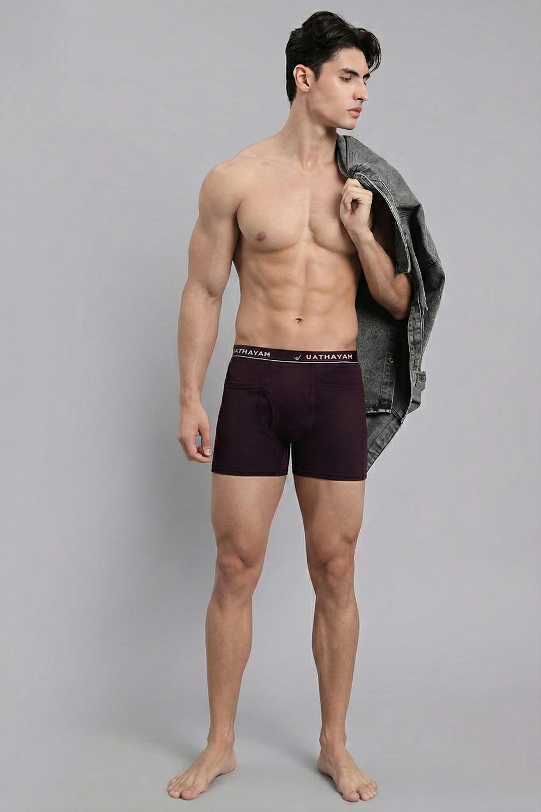 Man wearing dark red boxer briefs with a gray jacket over his shoulder on a gray background