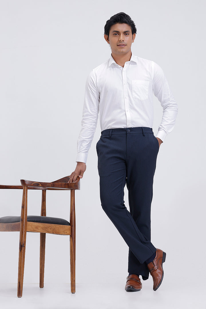 Man wearing a white shirt and navy pants standing next to a wooden chair on a white background