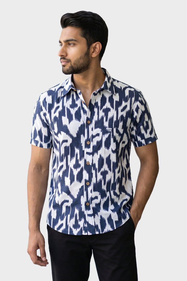 A man wearing a short-sleeve button-up shirt with a navy blue and white ikat pattern, paired with black trousers, standing against a light gray background.