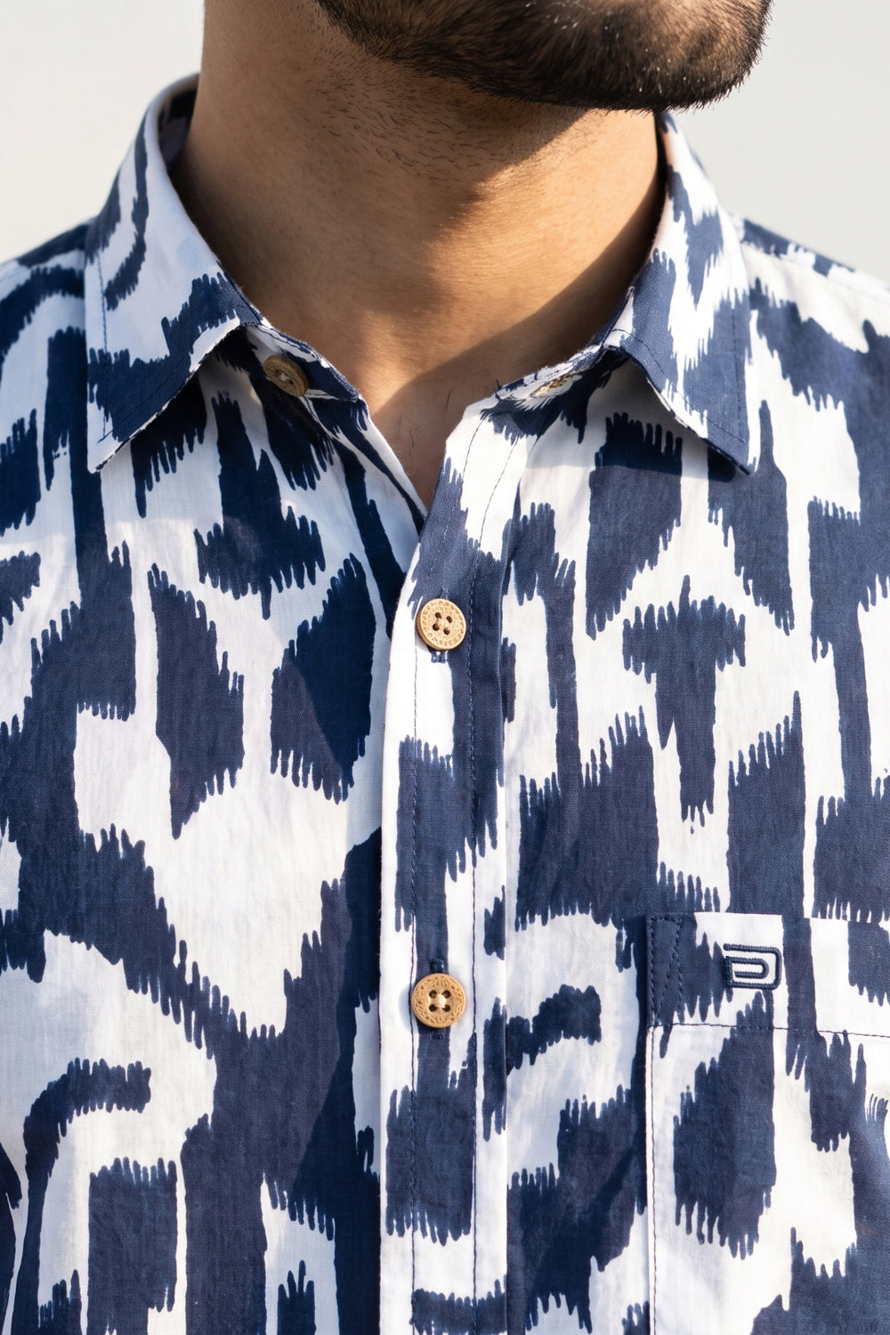 Close-up of a man wearing a navy and white ikat patterned button-down shirt with brown buttons and a chest pocket featuring a small logo.