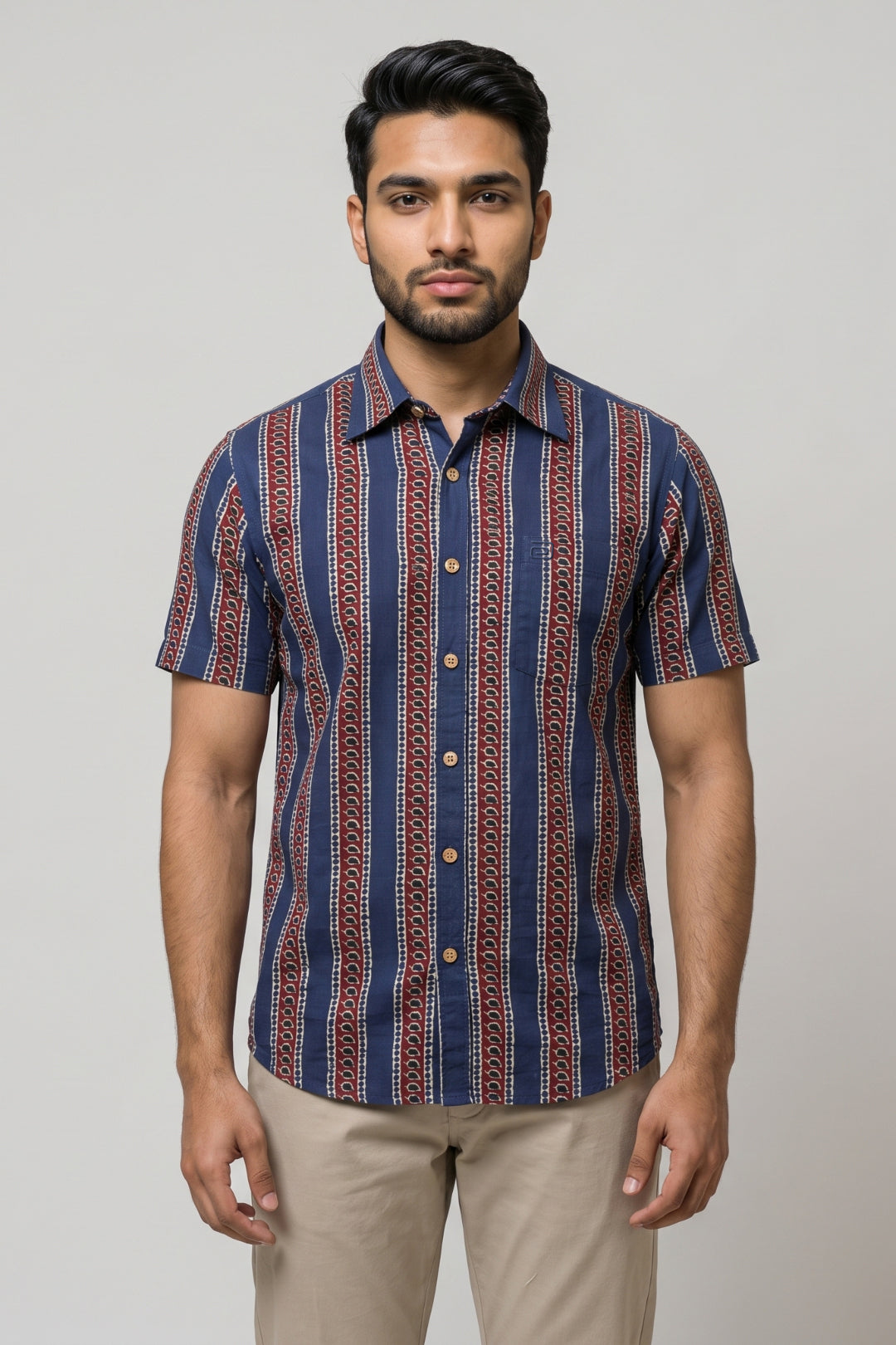 A man wearing a short-sleeve button-up shirt with vertical navy, red, and beige patterns, paired with beige trousers, standing against a light gray studio background.