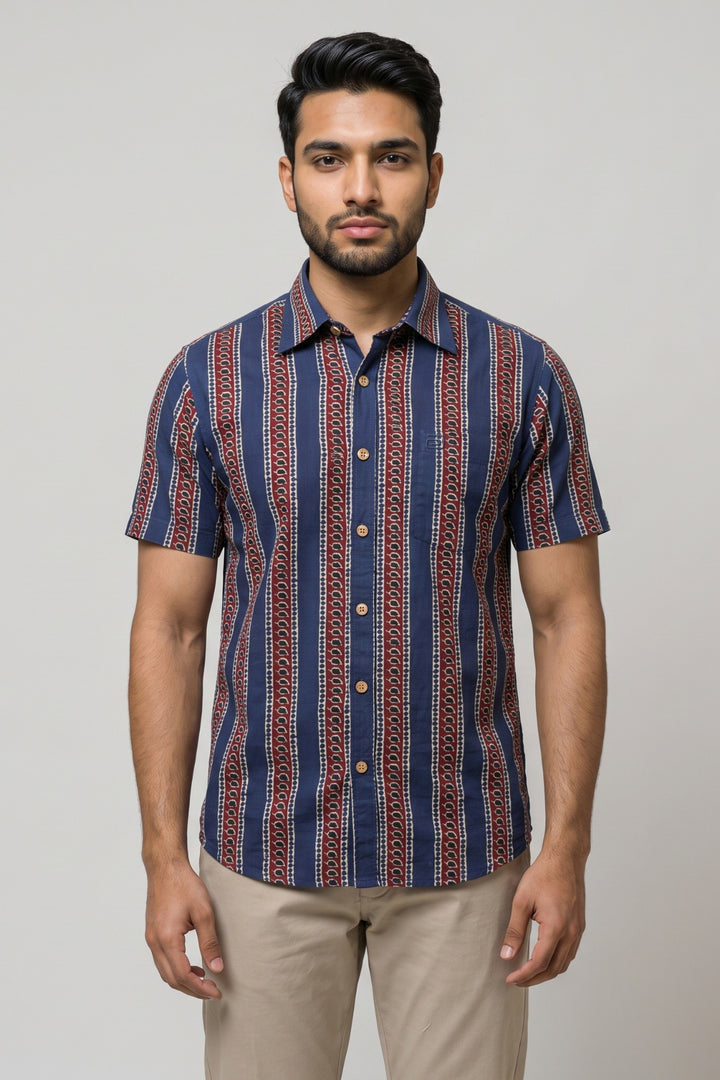 A man wearing a short-sleeve button-up shirt with vertical navy, red, and beige patterns, paired with beige trousers, standing against a light gray studio background.