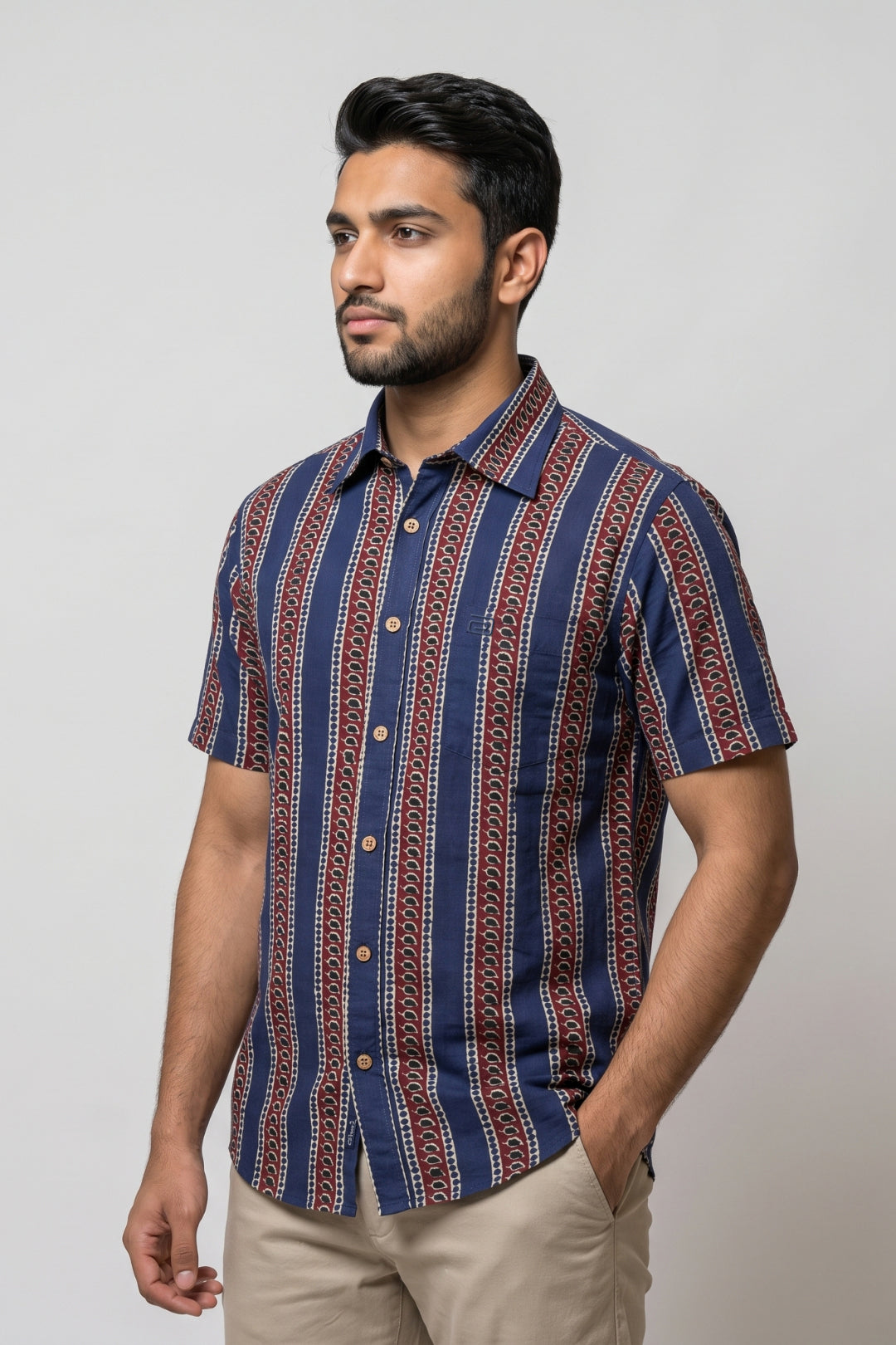 A man wearing a short-sleeve button-up shirt with vertical navy blue, red, and beige geometric stripes, paired with khaki pants, standing against a light gray studio background.