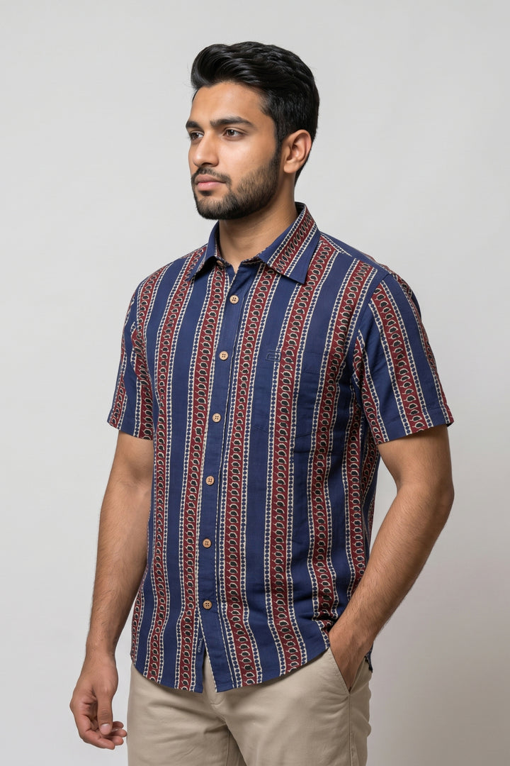 A man wearing a short-sleeve button-up shirt with vertical navy blue, red, and beige geometric stripes, paired with khaki pants, standing against a light gray studio background.