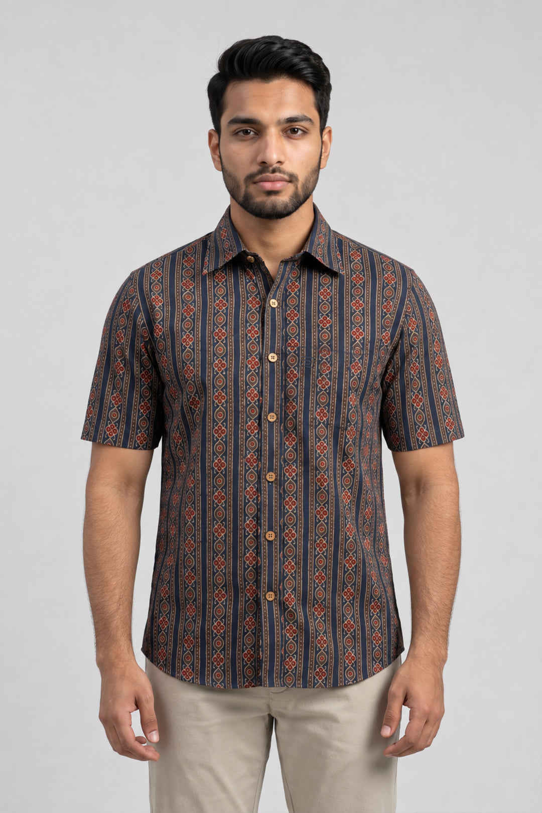 A man wearing a short-sleeve button-up shirt with a vertical geometric pattern in dark blue, rust orange, and beige, paired with beige trousers, against a light gray studio background.