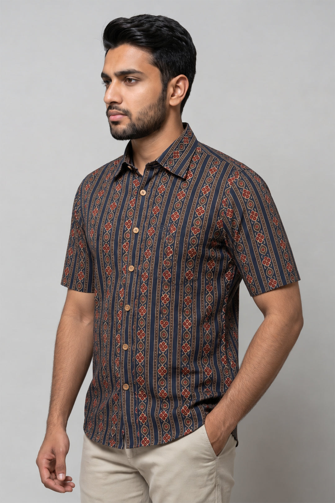 A man wearing a short-sleeve button-up shirt with a dark navy and brown geometric pattern, paired with beige trousers, standing against a solid gray studio background.