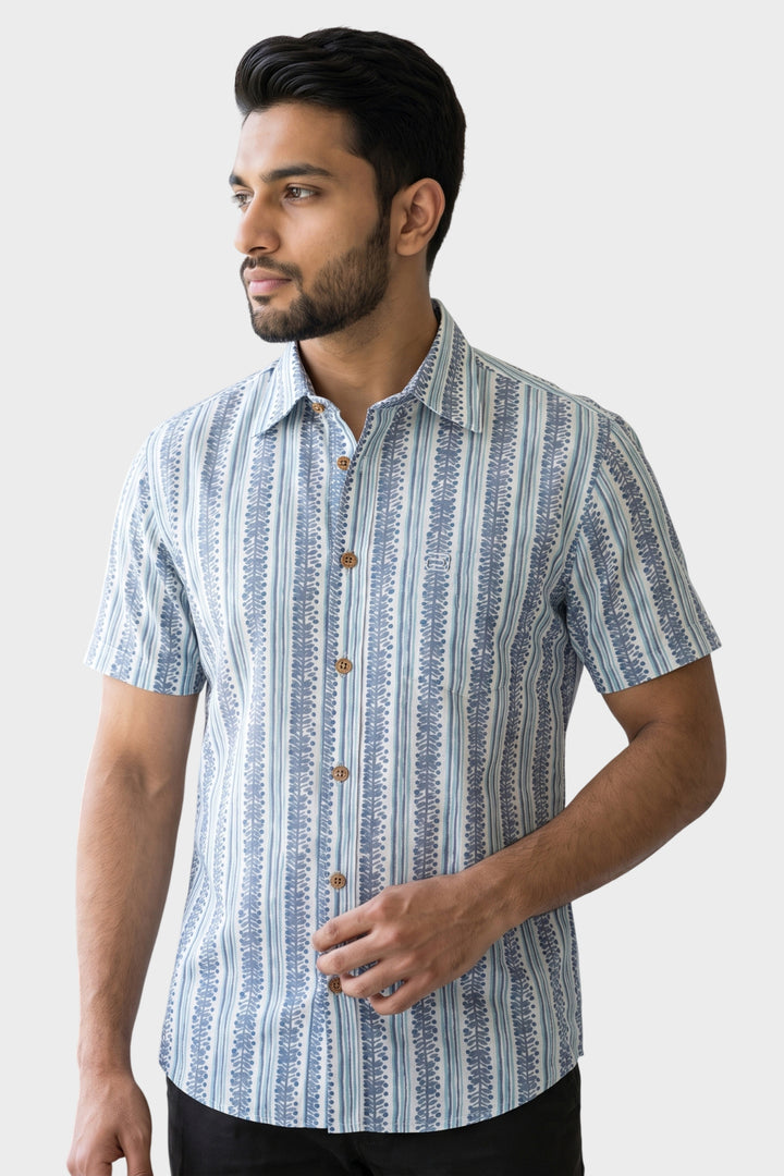 A man wearing a short-sleeve button-up shirt with a vertical blue and white pattern, posed against a light gray studio background.