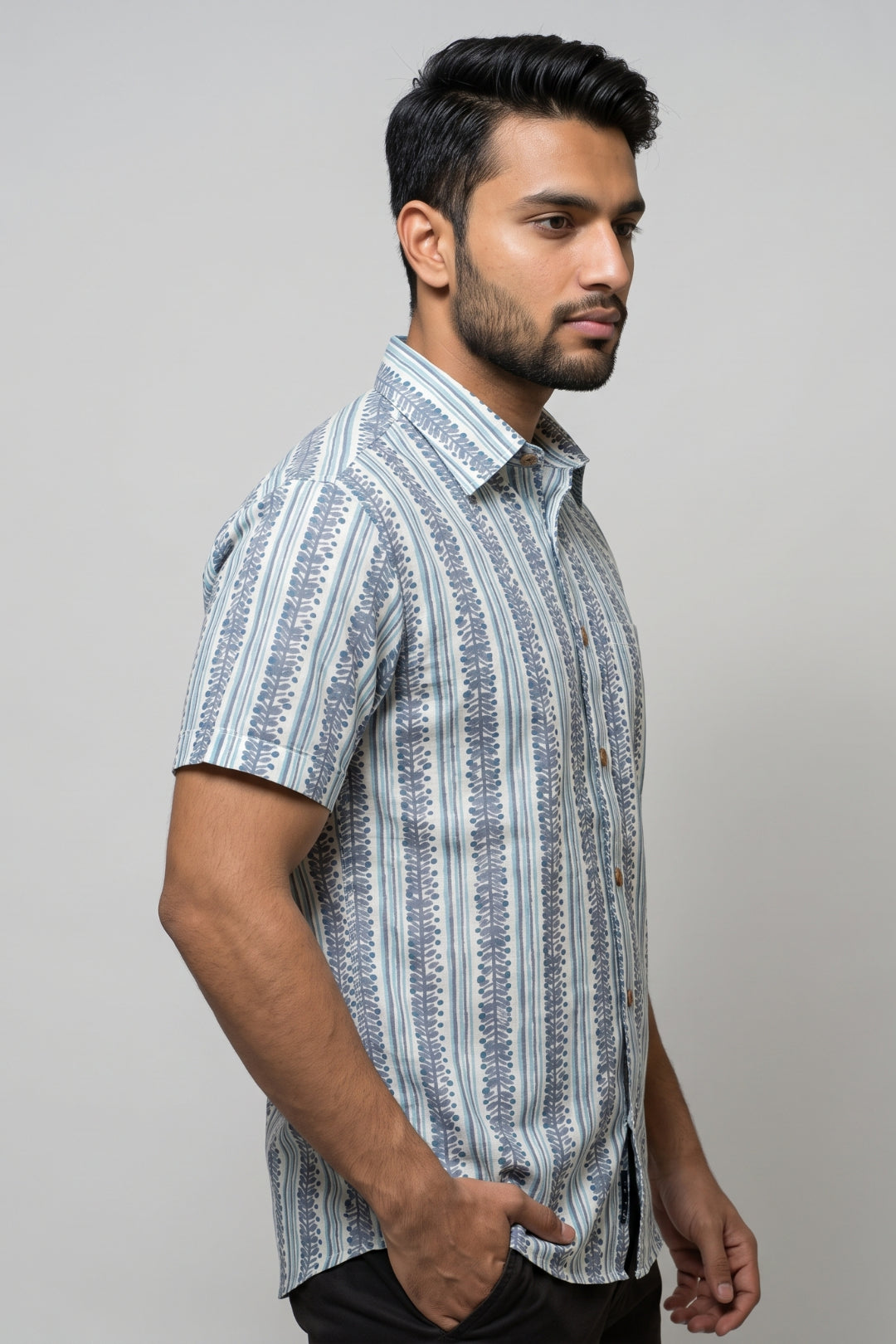 A man wearing a short-sleeve button-up shirt with vertical blue and white patterns, photographed against a plain light gray studio background.