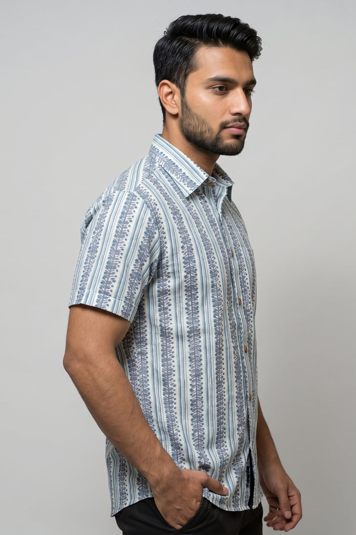A man wearing a short-sleeve button-up shirt with vertical blue and white patterns, photographed against a plain light gray studio background.