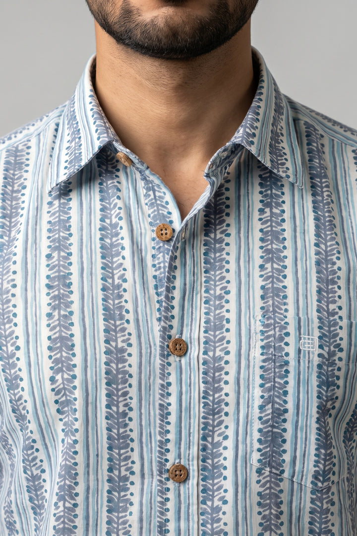 A close-up of a man wearing a light blue and white patterned button-down shirt with vertical leafy stripes and brown buttons.