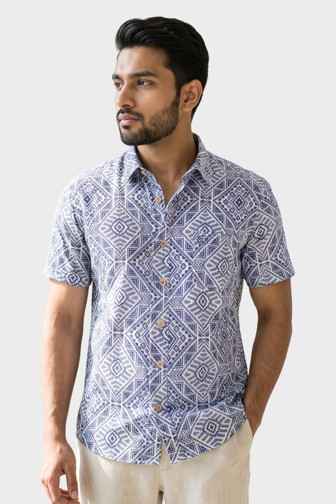 A man wearing a short-sleeve button-up shirt with a blue and white geometric pattern, paired with beige trousers, photographed against a light gray studio background.