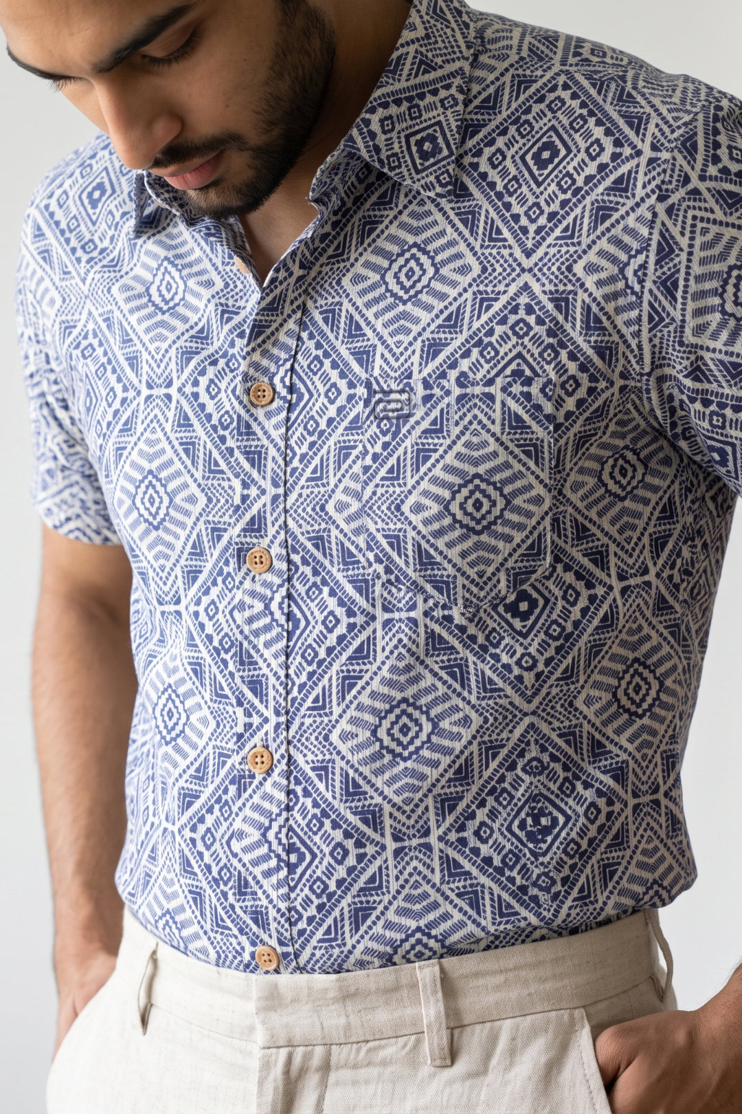 A man wearing a short-sleeve button-up shirt with a blue and white geometric pattern, paired with light beige trousers, shown from the chest down against a plain light background.