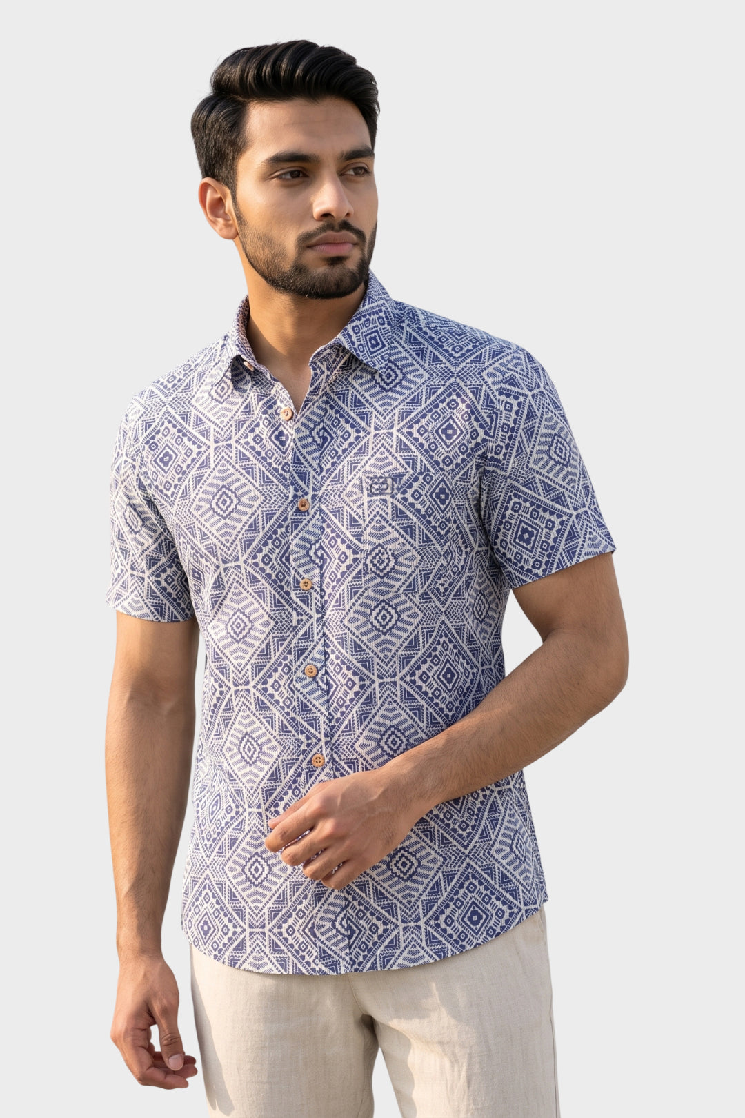 A man wearing a short-sleeve button-up shirt with a blue and white geometric pattern, paired with light beige trousers, set against a solid light gray background.