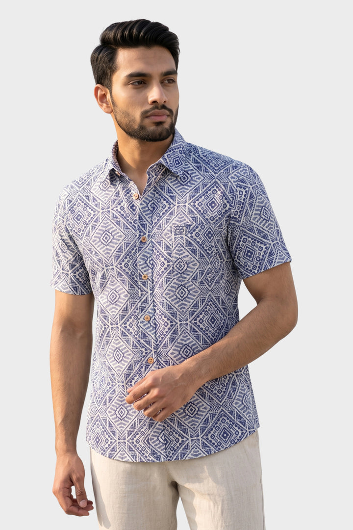 A man wearing a short-sleeve button-up shirt with a blue and white geometric pattern, paired with light beige trousers, set against a solid light gray background.