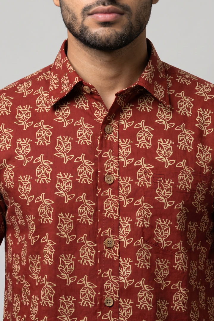 A close-up of a man wearing a rust-red button-down shirt with a beige floral block print pattern. The image is cropped from the lower face to the chest, showing the collar, front buttons, and fabric texture against a neutral gray background.