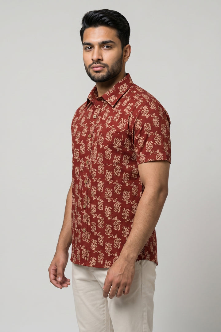 A man wearing a short-sleeve button-up shirt with a rust red and beige floral block print, paired with off-white trousers, standing against a light gray studio background.