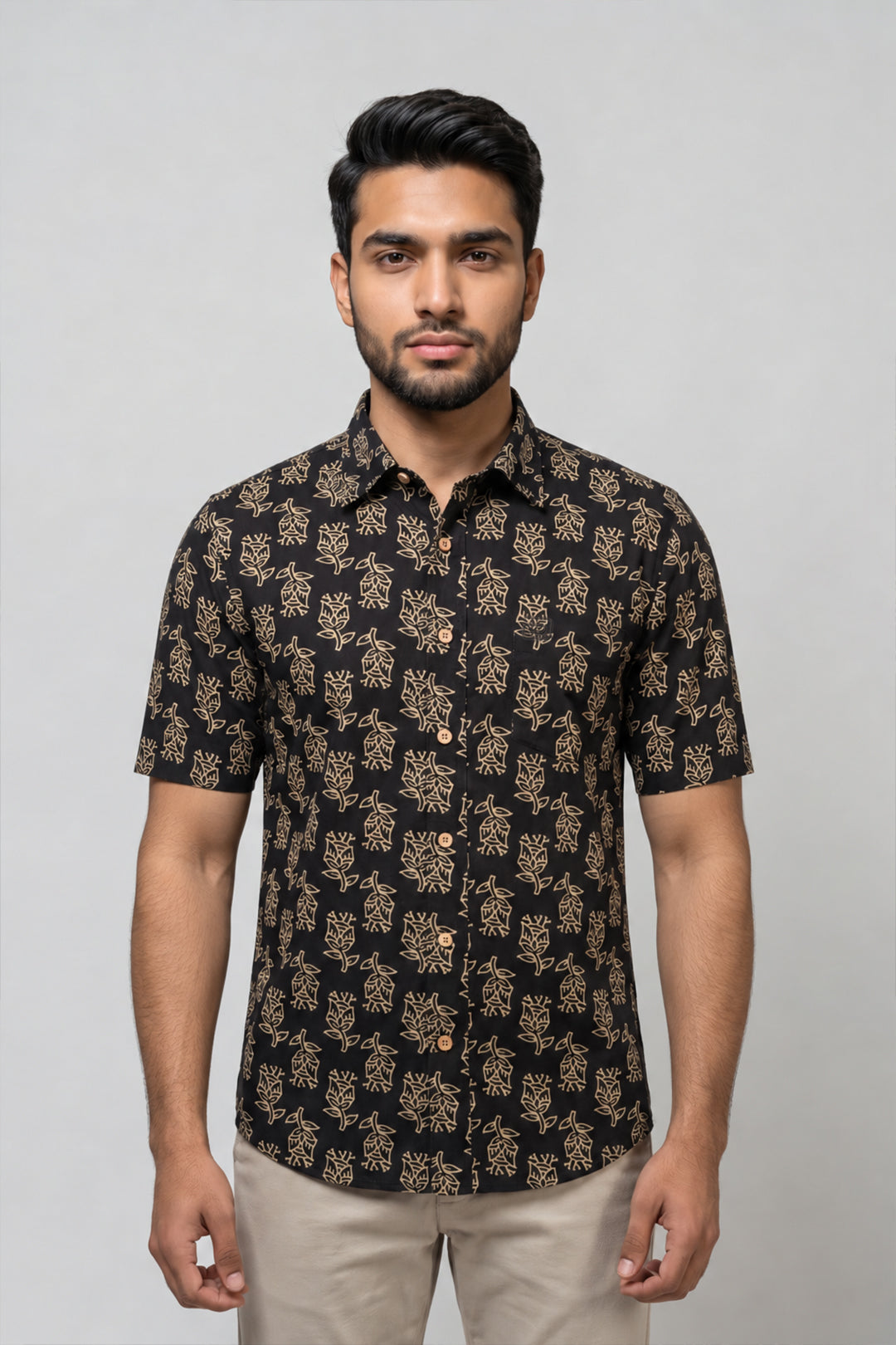 A man wearing a short-sleeve button-up shirt with a black base and beige floral pattern, paired with beige trousers, standing against a light gray studio background.