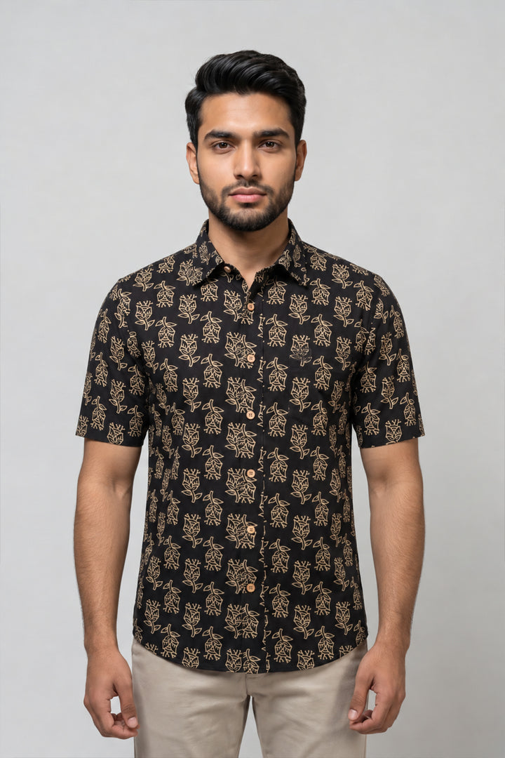 A man wearing a short-sleeve button-up shirt with a black base and beige floral pattern, paired with beige trousers, standing against a light gray studio background.
